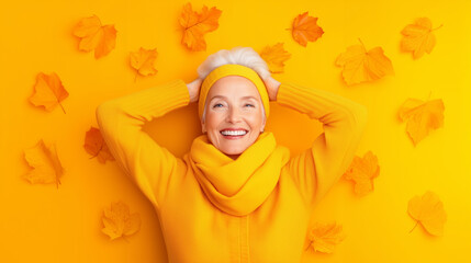 A woman smiles brightly while lying on a yellow background scattered with autumn leaves. Dressed in a cozy yellow sweater and scarf, she embodies the warmth of the fall season
