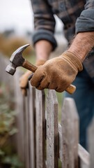 Man in work gloves repairing wooden fence with hammer, closeup on hands and tool in outdoor setting
