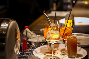 Colorful cocktails with citrus slices and straws served on trays in a dimly lit bar. Glasses filled with Aperol Spritz and iced drinks ready for guests on a vibrant evening.
