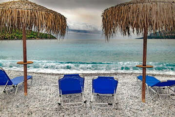 straw umbrella on a beach of white pebbles  with lounge chairs in front of a beautiful sea under cloudy sky in Peloponnese, Greece