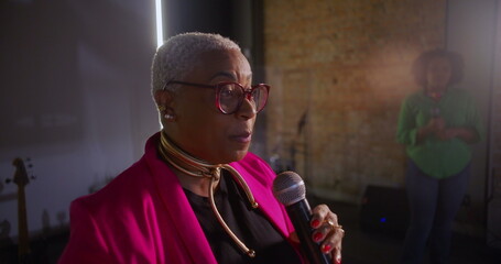 Close-up of a female speaker passionately delivering a sermon, holding a microphone with focus on expression and connection, brick wall and attendee in blurred background