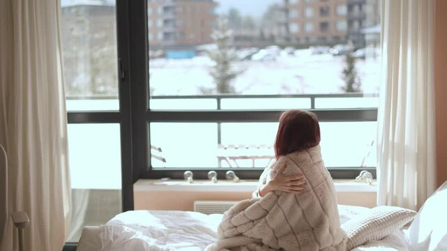 On a chilly winter morning, a woman wrapped in a cozy blanket gazes at the serene snowy landscape outside her window. She reflects on her thoughts while staying warm and enjoying the stillness