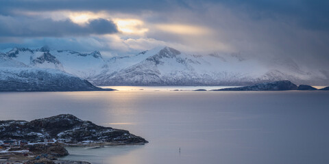 mountains and valleys formed by glaciers in winter, changing weather moods on the fjord between steep, snow-covered mountains in Troms, Norway. blue sky between dark clouds on the sky, snowy mountains
