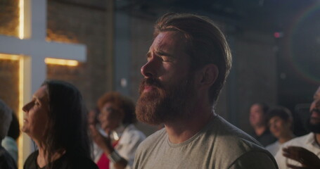 Bearded man praying during church worship, hands raised in devotion, illuminated cross and diverse congregation in background, expression of faith and spirituality