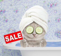 Funny kitten with towel on it head and with slices of cucumber on it eyes takes a bath at home, shows thumbs up gesture and signboard with labeled "sale"