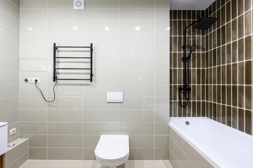 well-lit bathroom with beige tiles, a white toilet, a black towel warmer, and a bathtub with a...