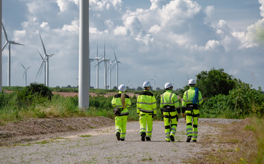 Maintenance engineer team standing at windmills at wind turbine farm. Group of people wear safety helmet and uniform working at alternative renewable energy wind station. Sustainable energy technology