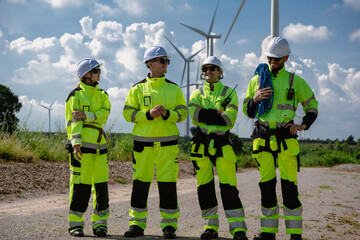 Maintenance engineer team standing at windmills at wind turbine farm. Group of people wear safety helmet and uniform working at alternative renewable energy wind station. Sustainable energy technology