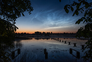Tranquil lake at twilight with lily pads, pier remnants, framed by leafy trees, and glowing noctilucent clouds in a vibrant evening sky,slilver cloude