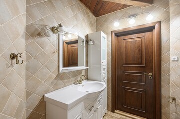 well-lit bathroom features beige diamond-patterned tiles, a white vanity with a sink, and a wooden door. Brass fixtures and a wooden ceiling beam add a rustic touch
