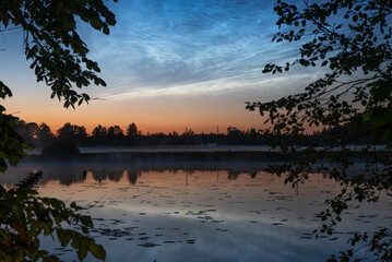 Tranquil lake at twilight with lily pads, pier remnants, framed by leafy trees, and glowing noctilucent clouds in a vibrant evening sky,slilver cloude