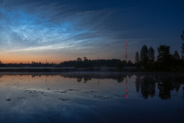Twilight view of a calm lake with reeds, pier stubs, noctilucent clouds, and a red-lit radio tower reflected in the water near forest edge,silver clouds