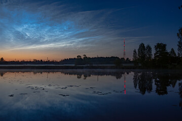 Twilight view of a calm lake with reeds, pier stubs, noctilucent clouds, and a red-lit radio tower reflected in the water near forest edge,silver clouds
