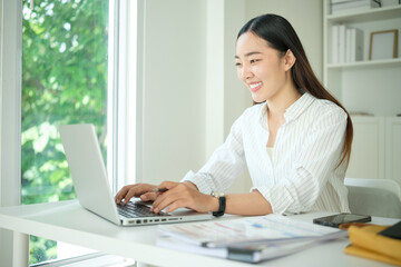 Smiling Asian woman in a white shirt working on a laptop at a bright modern desk near a window