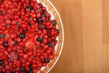 A vibrant mix of frozen red berries fills a decorative bowl, placed on a light wooden surface with ample copy space to the right