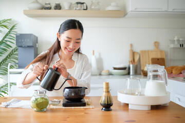 Young woman preparing matcha tea in a bright modern kitchen, pouring hot water into a traditional...