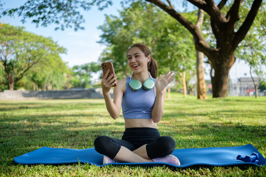 Young woman sitting cross-legged on a yoga mat in the park, smiling and waving during a video call with her smartphone, headphones around her neck.
