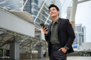 Successful businessman standing outdoors in the city, holding his phone and smiling.