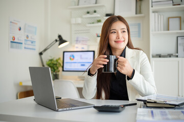 Confident young professional holding a black mug and smiling while sitting at her desk with laptop and documents.