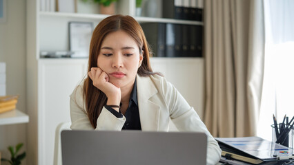 Young Asian businesswoman appearing fatigued and stressed while working on a laptop at the office desk.