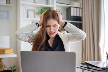 Young Asian businesswoman clutches her head in frustration while staring at a laptop screen at her office desk.