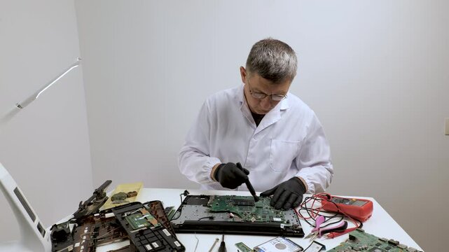 Technician cleaning motherboard, Man in white lab coat and black gloves repairing and inspecting a disassembled laptop motherboard with multimeter and brush tools on a clean white table