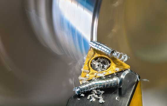 Closeup of lathe tool bit with metal shavings curled up spiral at turning rotating workpiece. Sharp blade cutter with carbide insert and rolled up swarf in abstract motion detail with blur background.