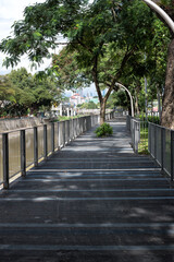 Scenic Riverside Walkway in Brickfields, Kuala Lumpur. The pathway runs parallel to the river, with tall buildings and tropical trees in the background.
