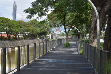 Fototapeta premium Scenic Riverside Walkway in Brickfields, Kuala Lumpur, Malaysia. The pathway runs parallel to the river, with tall buildings and tropical trees in the background.