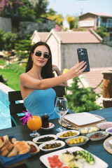 A 20s brunette in blue dress and sunglasses snaps a selfie at the breakfast table with the hotel...
