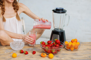 Woman pouring fresh homemade strawberry smoothie into a glass from blender cup on rustic wooden table with bowls of strawberries, apricots, and ice. Healthy summer drink concept.