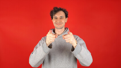 A happy, smiling curly-haired man in a gray hoodie points his index fingers in front of him....