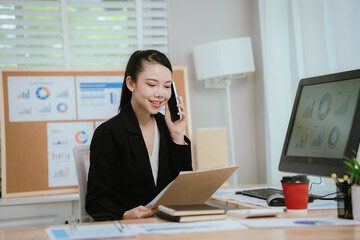 Obraz premium Confident Asian businesswoman smiling at her desk with laptop and financial charts, planning strategy and analyzing data for business success.