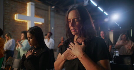Women in prayer during church service, hands on chest in devotion, illuminated cross in the background, community gathering in spiritual reflection and worship