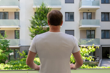 A man in a white t-shirt stands on a balcony looking out over an apartment block, enjoying the view and a moment of solitude. © Ernest