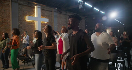 Multicultural congregation clapping and smiling during church worship, illuminated cross in...