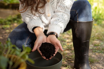 Close-up view of a person wearing black rubber gardening boots standing next to a flower pot and a watering can in an outdoor garden. The image evokes a peaceful gardening moment, with natural light.