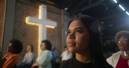 Young woman sitting in church during worship service, gazing forward with thoughtful expression, illuminated cross and diverse congregation in background
