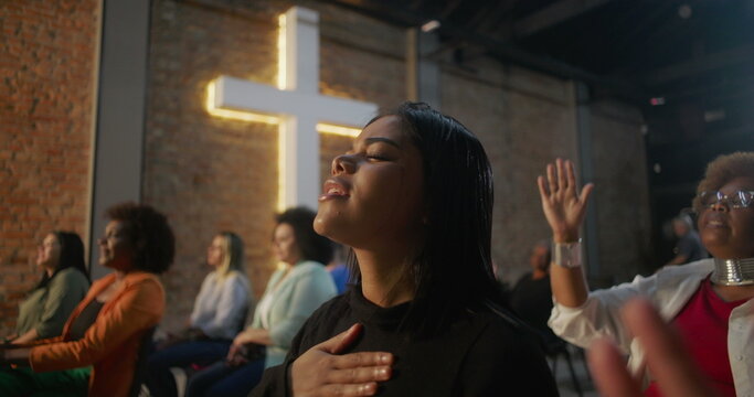 Women singing in prayer during church service, one raising hand in worship while another holds chest in devotion, illuminated cross and congregation in background