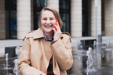 Fototapeta premium A fifty-year-old smiling woman talks on her mobile phone next to a fountain in the city in autumn.