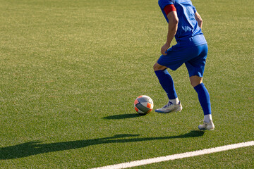 Soccer player in blue uniform dribbling the ball along the sideline of a football field during a sunny day.