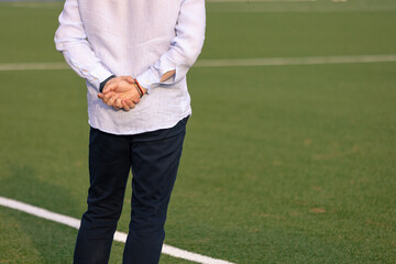 Back view of a man in formal casual attire standing on the football field with hands behind his back, possibly a coach or manager.