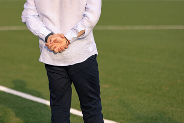 Back view of a man in formal casual attire standing on the football field with hands behind his back, possibly a coach or manager.