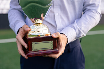 Man in a striped shirt holding a football trophy with a blank plaque on a soccer field.