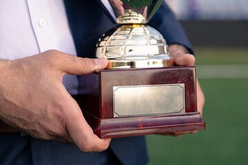 Close-up of a man in a suit holding a shiny football trophy with a blank plaque, standing on a soccer field.