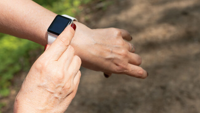Close-up of a woman using a smartwatch with a white strap in a green forest, tracking activity or checking notifications during an outdoor walk.