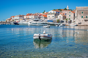 A picturesque coastal town of Primosten in Croatia features clear turquoise water, moored yachts, stone houses with red rooftops, and a peaceful Mediterranean atmosphere under a bright blue sky.