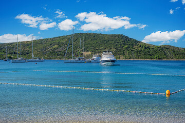 A tranquil coastal scene featuring clear turquoise waters, anchored yachts, and a green hillside under a bright blue sky in Primosten, Croatia.