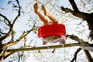 Closeup of feet of little toddler girl having fun on swing in domestic garden. Small child swinging under blooming trees on sunny day. Active leisure and activity outdoors