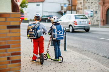 Two school kid boys in safety helmet riding with scooter in the city with backpack on sunny day. Happy children in colorful clothes biking on way to school.